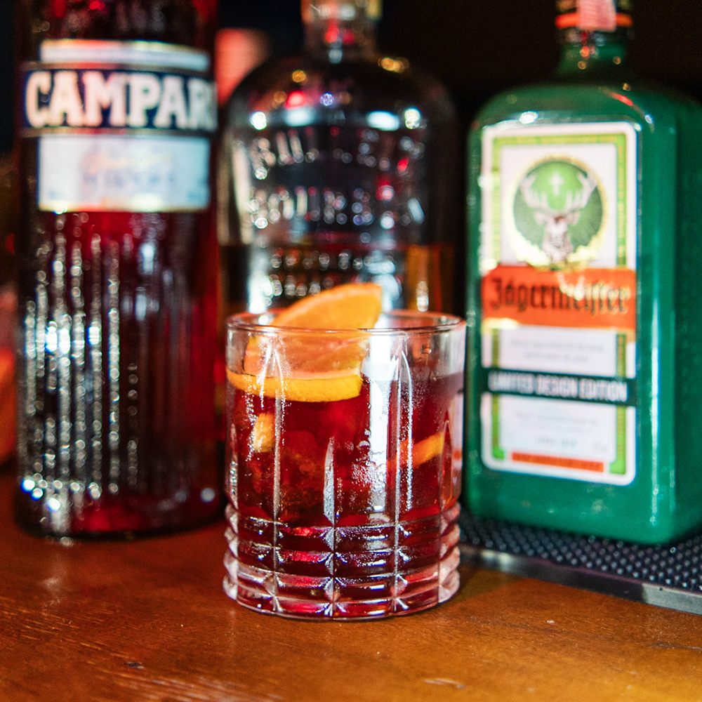 Drink com laranja no balcão, entre garrafas de Campari e Jägermeister